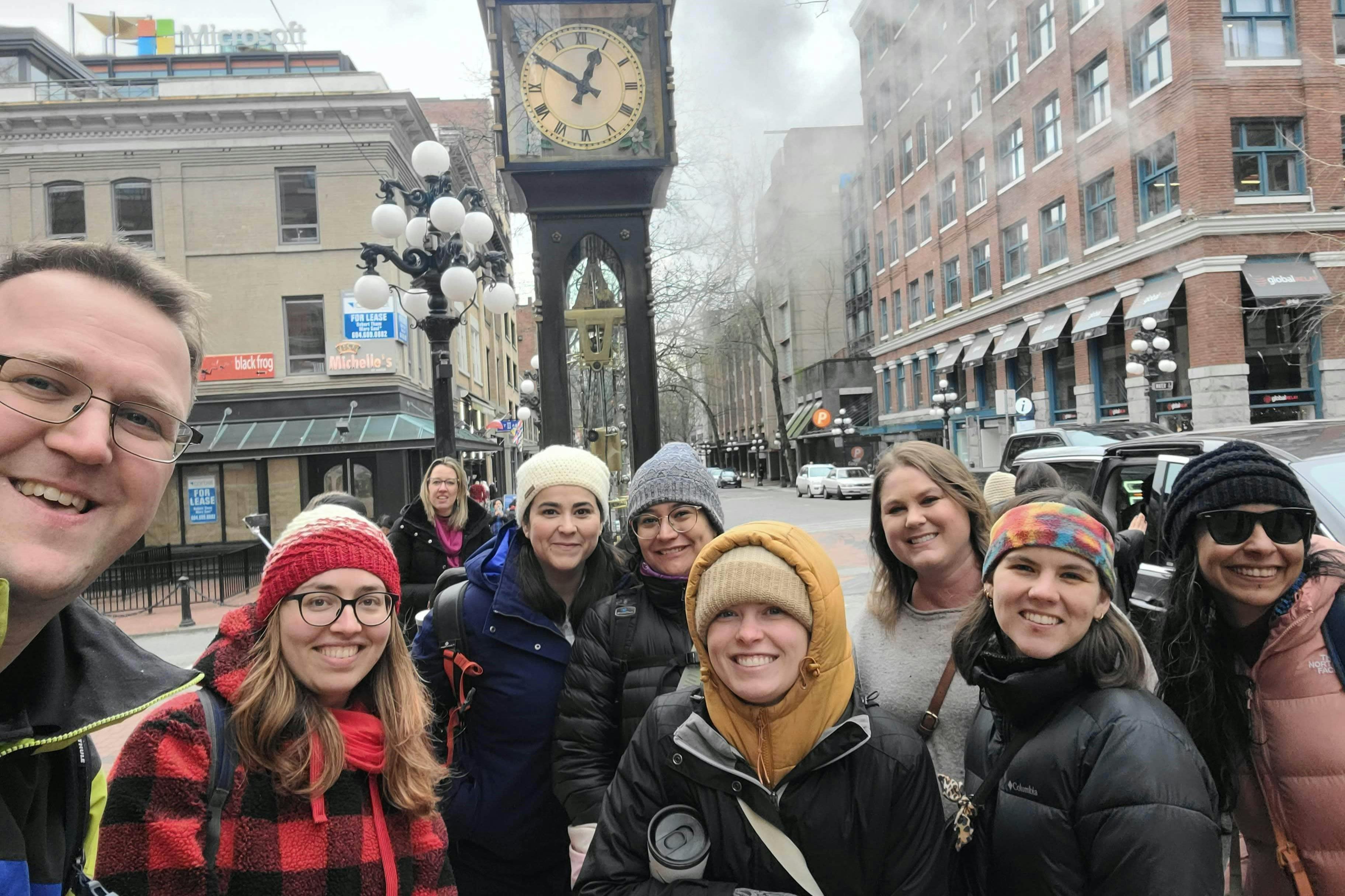 Skupina lidí, kteří se usmívají před parními hodinami Gastown v městské ulici s budovami a viditelnou lampou.