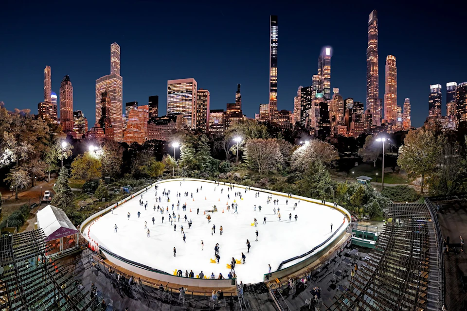 People are ice skating on a large outdoor rink illuminated by lights, surrounded by trees and modern city buildings at night.