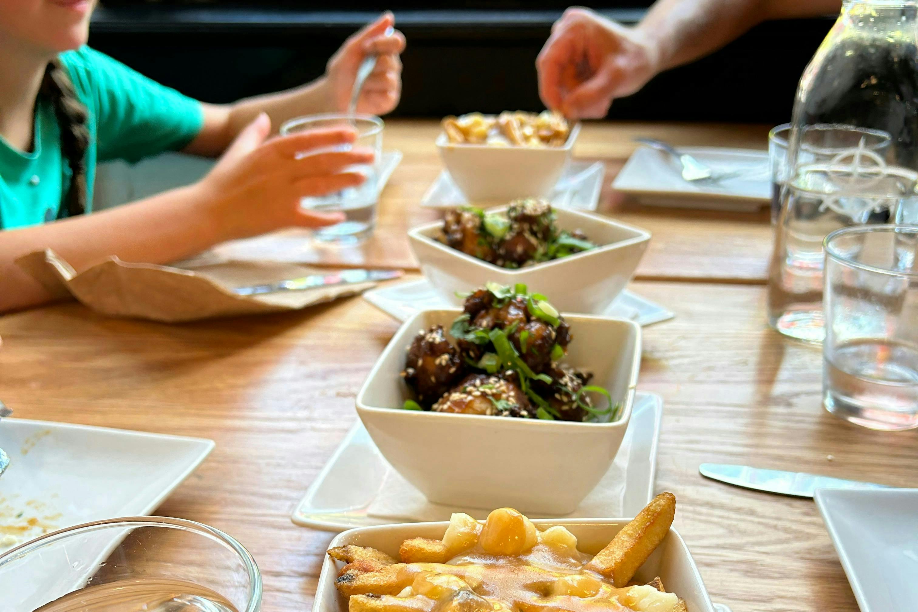 Un tavolo con ciotole di cibo, tra cui poutine e piatti guarniti con cipolle verdi, con mani che afferrano gli oggetti.
