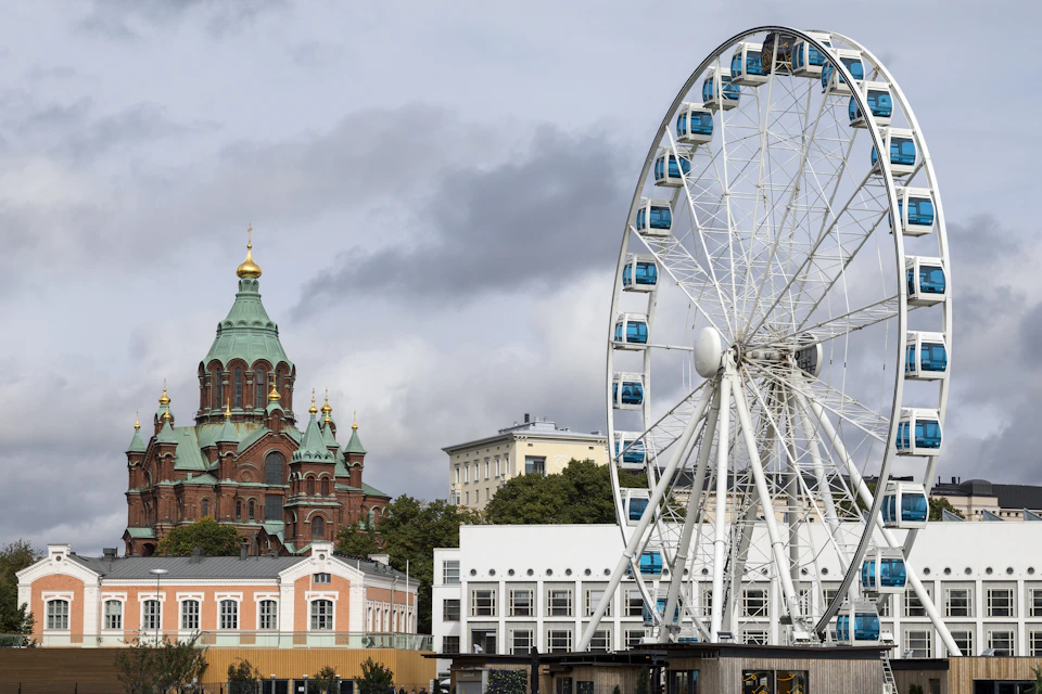 A Ferris wheel with blue gondolas stands in the foreground, while a red-brick church with green domes is visible in the background.