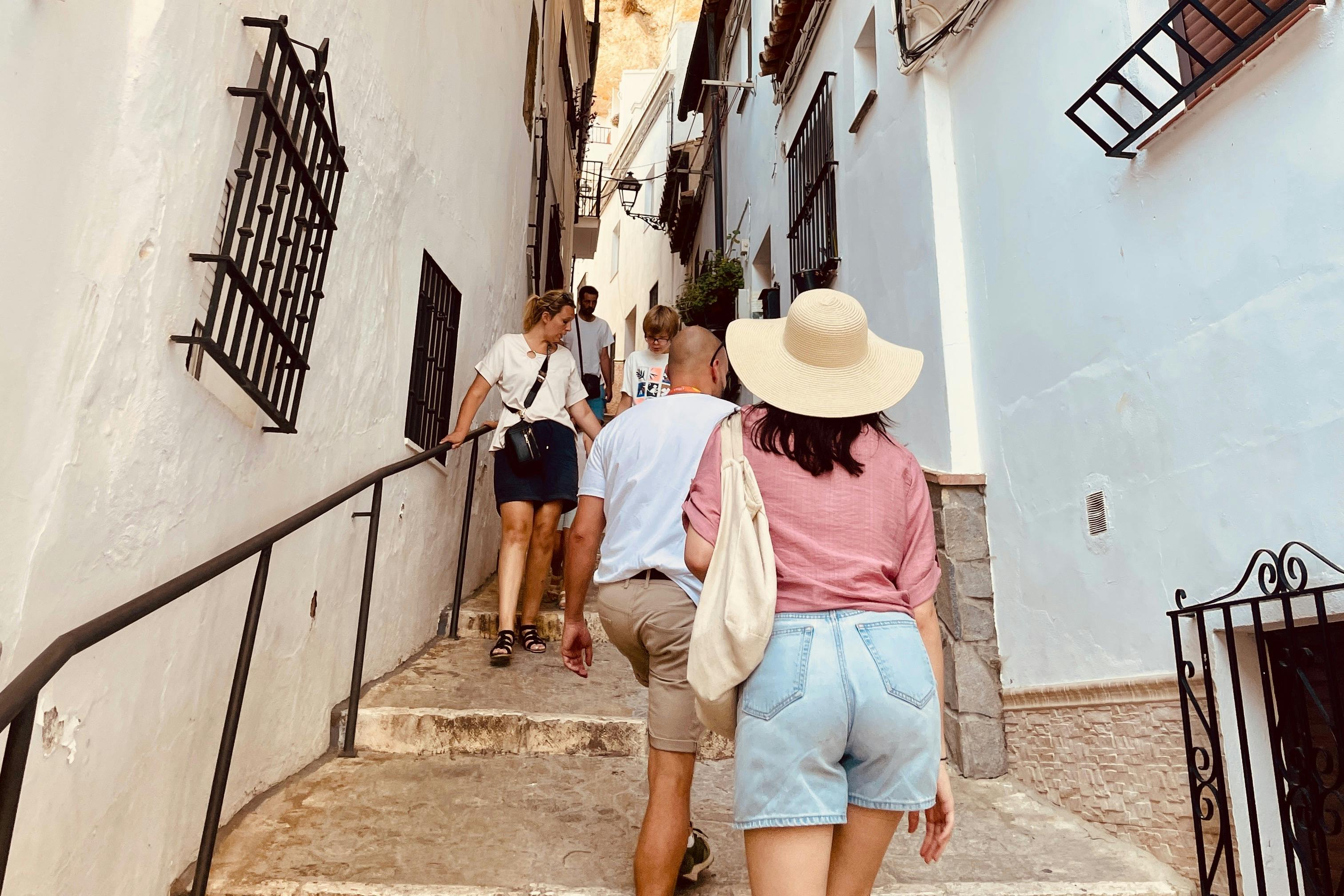 Άνθρωποι περπατούν σε ένα στενό δρομάκι με λευκά κτίρια και σιδερένια κάγκελα παραθύρων μια ηλιόλουστη μέρα.