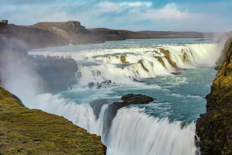 A broad waterfall with multiple tiers cascading over rocks, with people standing on a viewing platform in the distance.