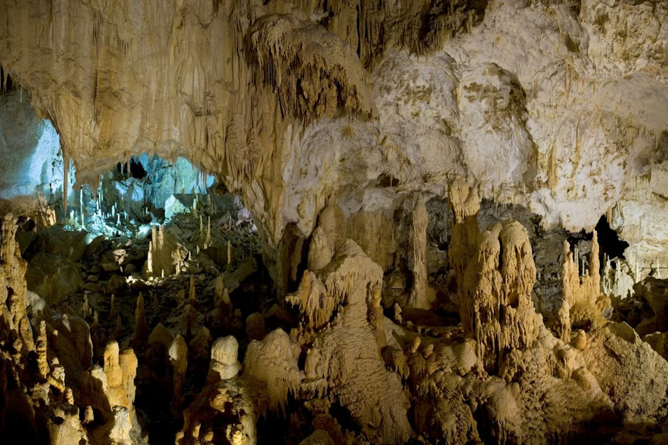 A cave interior with numerous stalactites and stalagmites, illuminated with soft natural and artificial lighting.