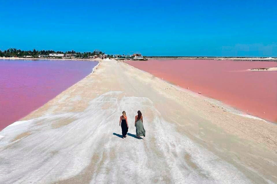 Two women walk on a sandy path between two vibrant pink lakes under a clear blue sky.