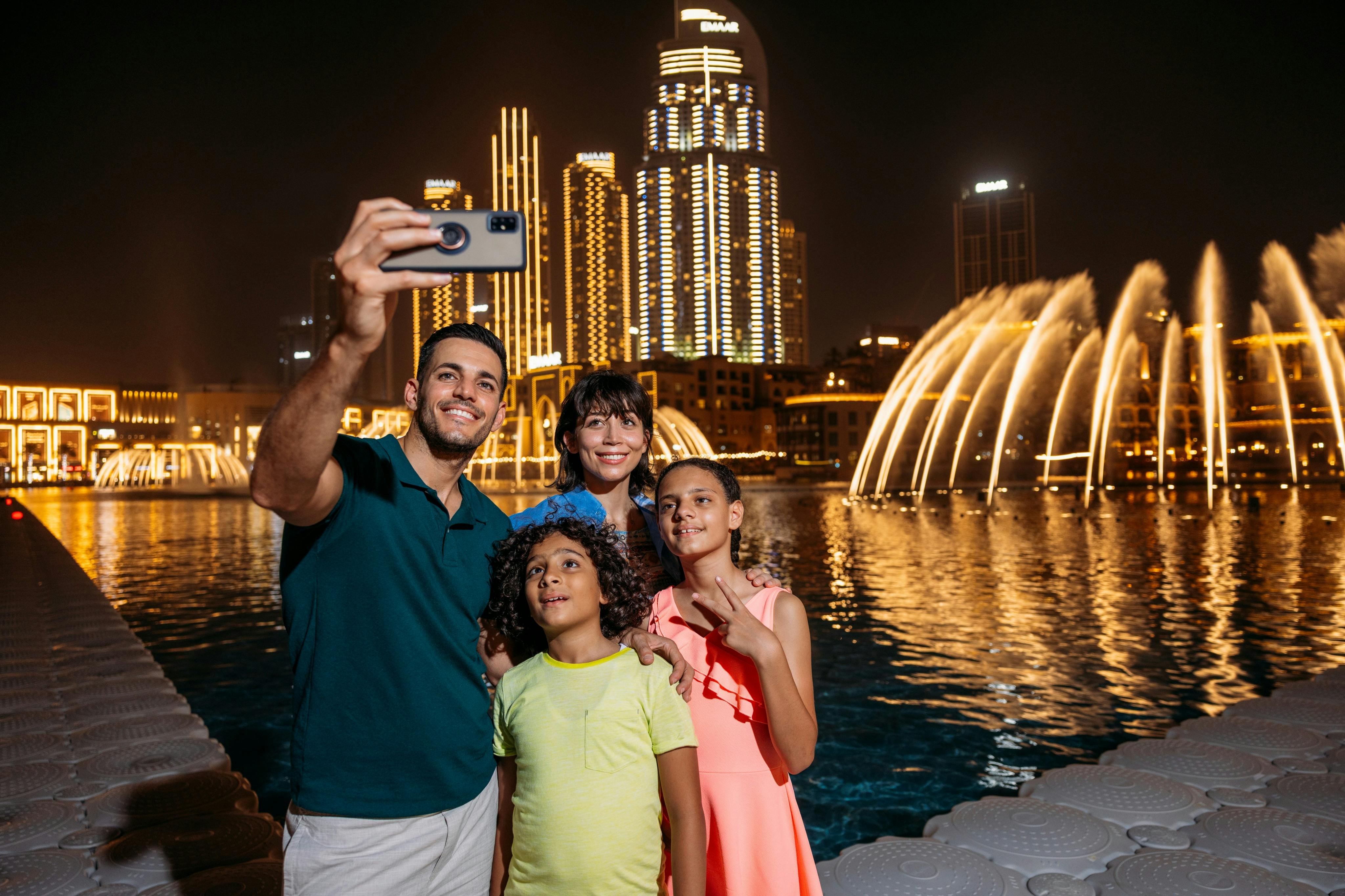 A family of four smiles while taking a selfie at night near illuminated fountains and tall buildings.