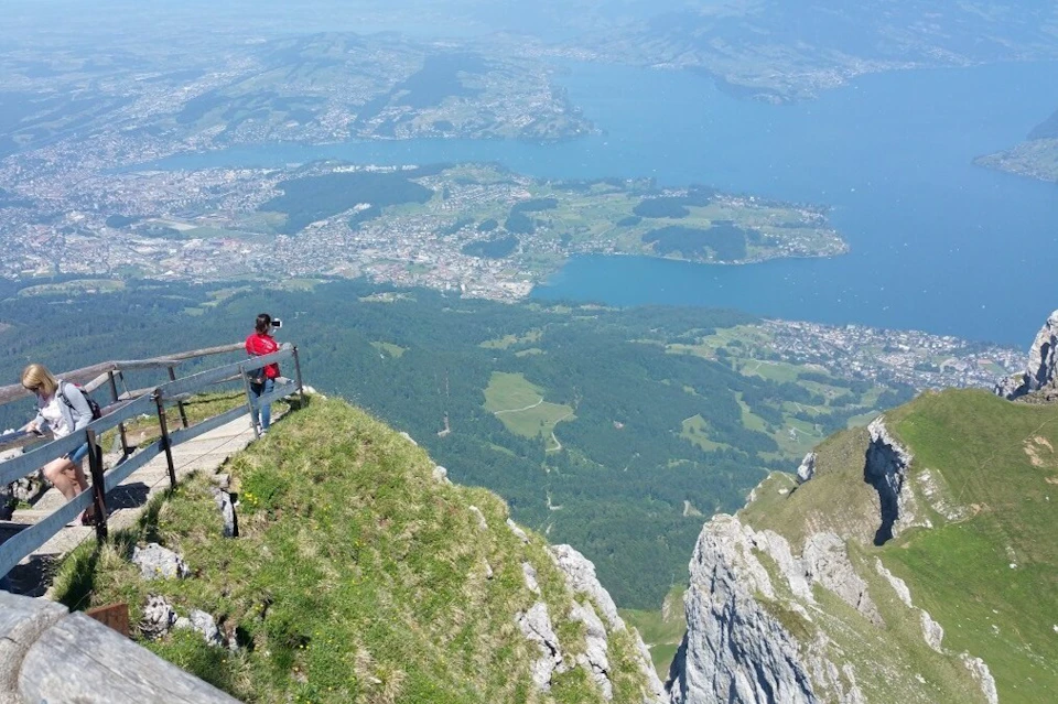 A person in a red jacket taking photos from a mountain viewpoint overlooking a vast landscape with a lake and a town.