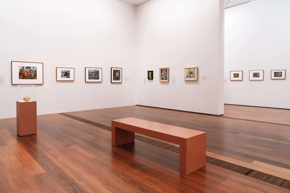 An art gallery with framed artwork on white walls and a wooden bench on a polished wooden floor.