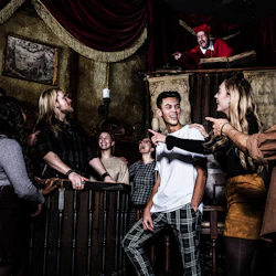 En grupp människor skrattar och pekar på en ung man som står framför dem, medan en man i en medeltida kostym gestikulerar från en balkong.