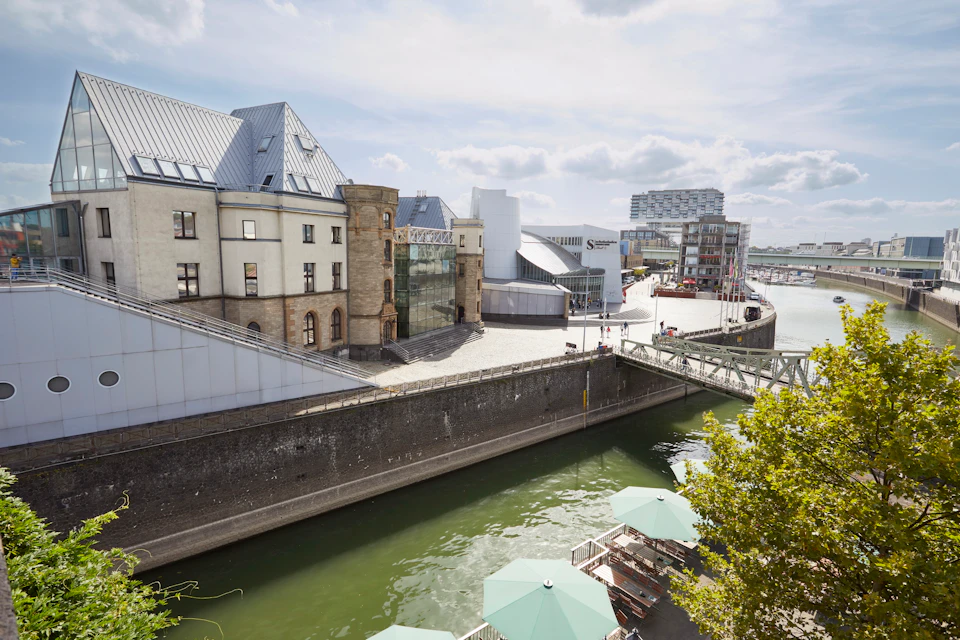 Modern buildings along a canal with green umbrellas on a deck in the foreground and a bridge crossing the water.
