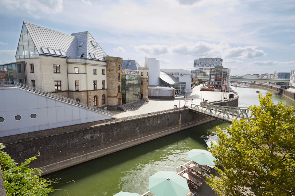 Modern buildings along a canal with green umbrellas on a deck in the foreground and a bridge crossing the water.