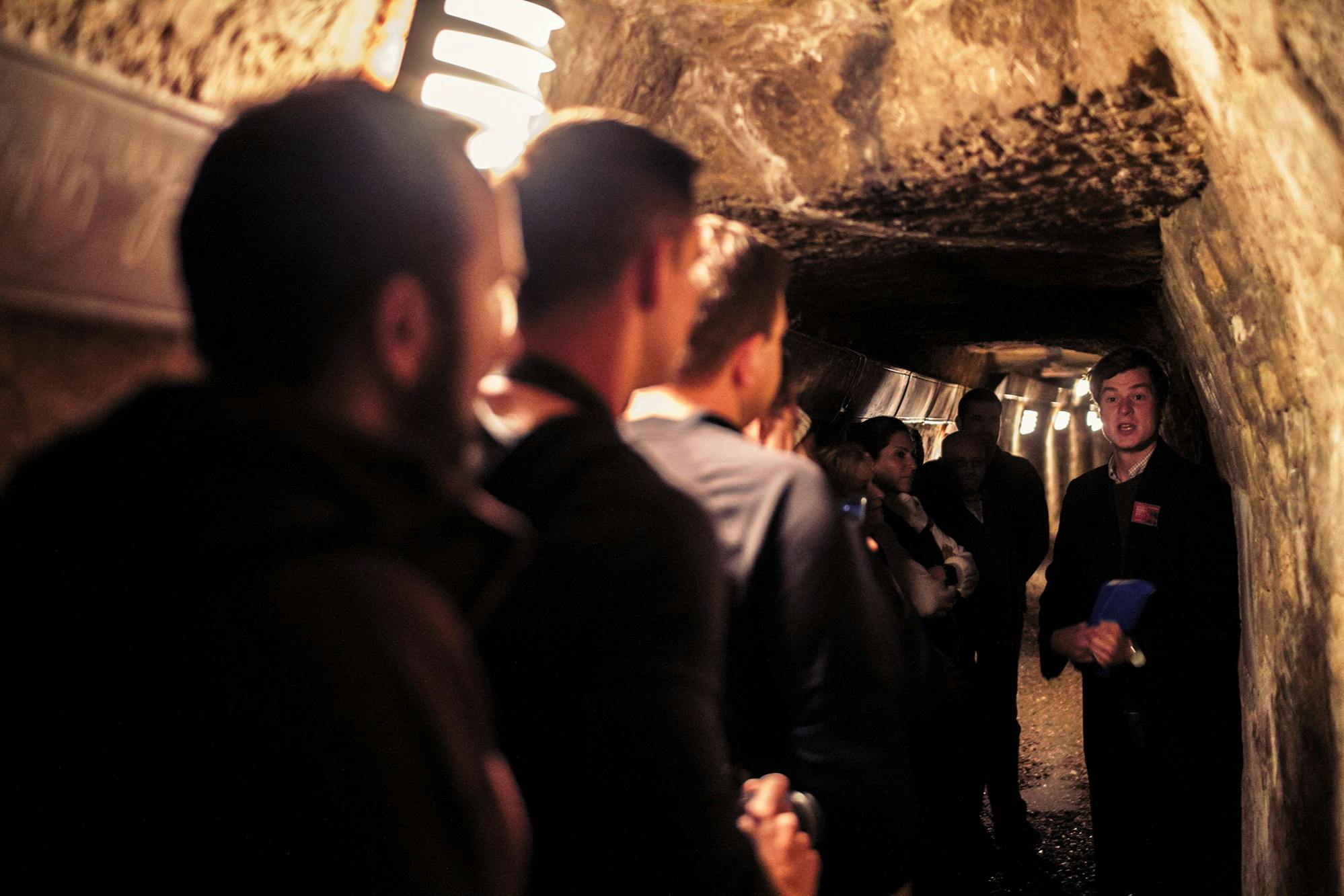 A group of people in a narrow, dimly lit underground tunnel, with a guide speaking to them holding notes.