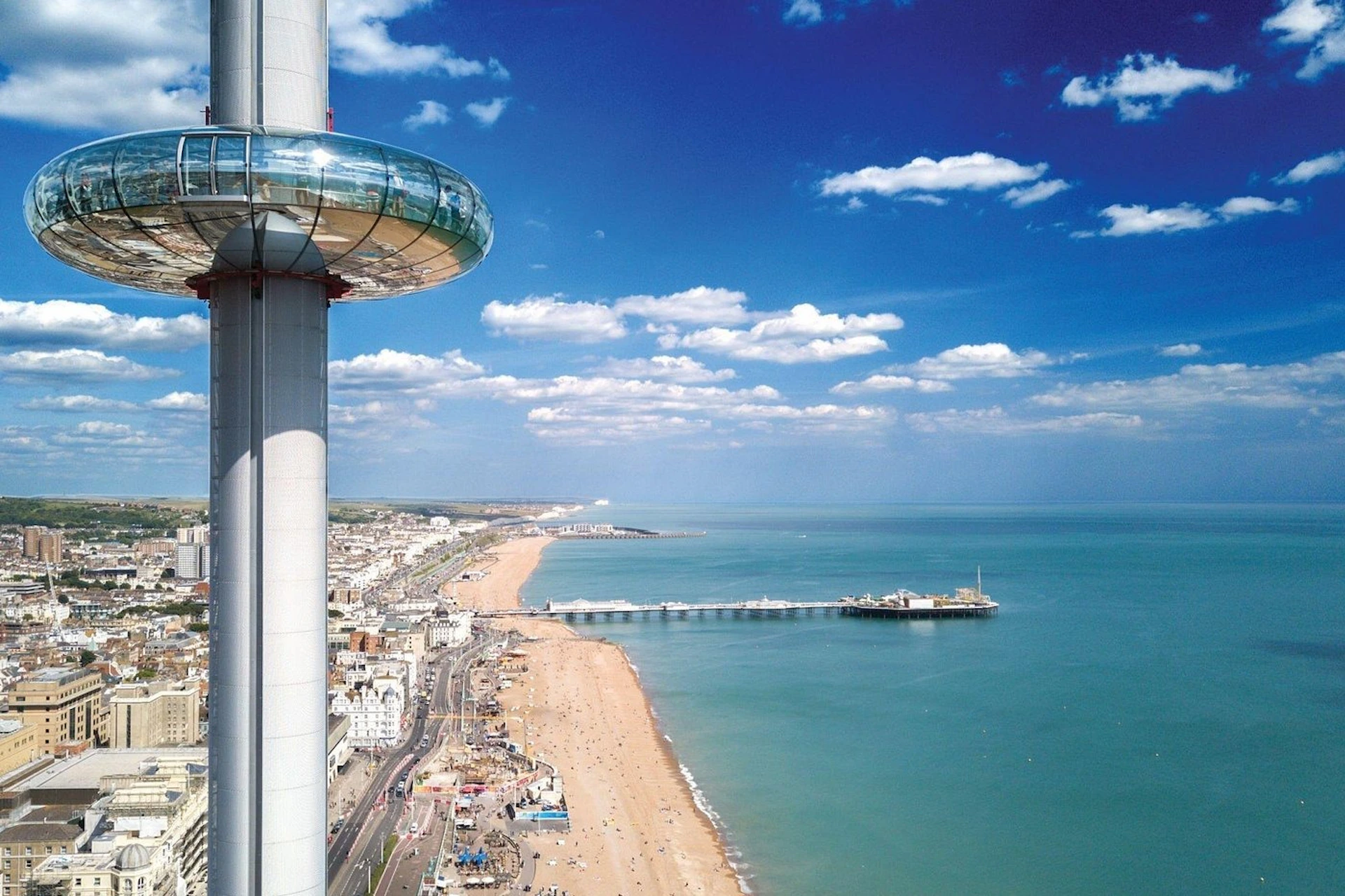 A seaside cityscape with a beach, pier, and a tall observation tower under a blue sky with clouds.