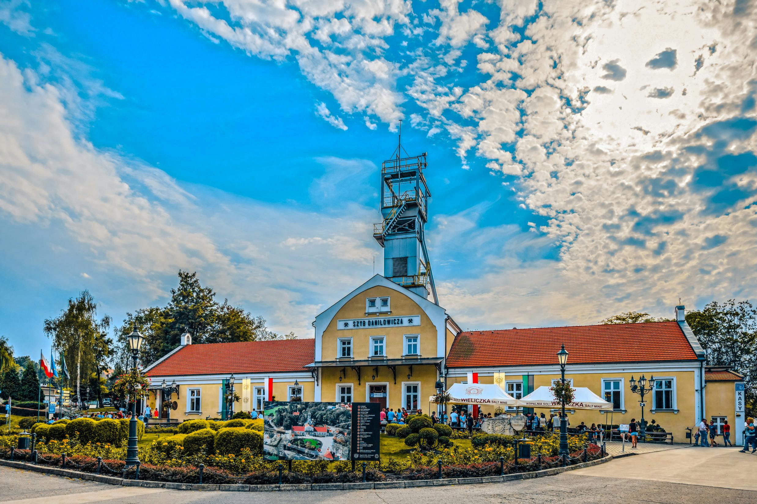 这座历史悠久的黄色建筑有一个红色的屋顶和一座塔楼，周围环绕着人群和绿色植物，在一片多云的天空下。