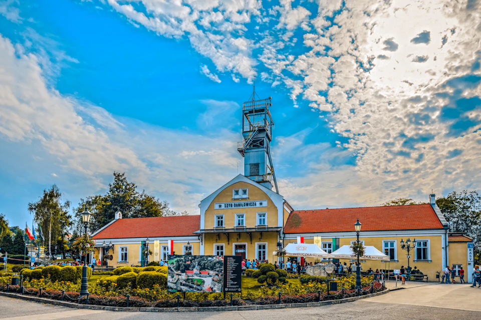 A historic yellow building with a red roof and a tower, surrounded by people and greenery, under a partly cloudy sky.