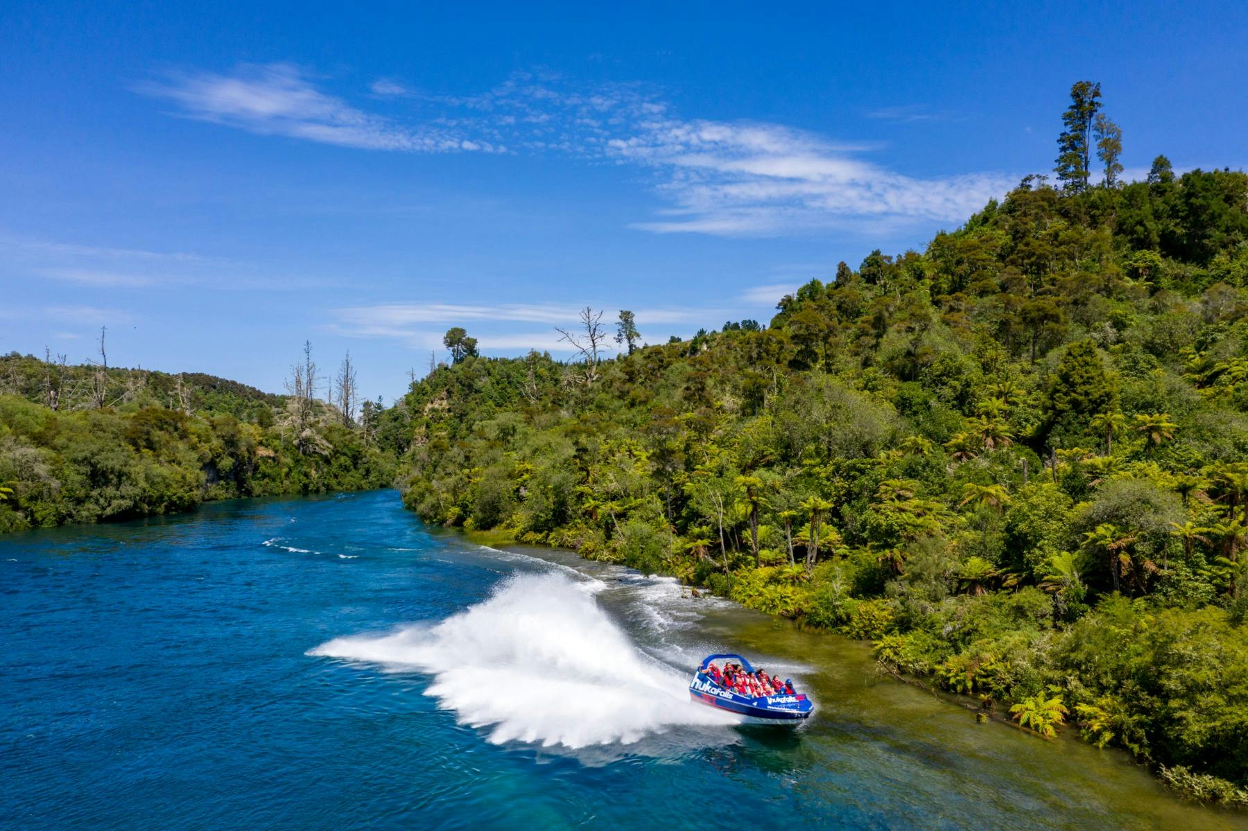 A speedboat with passengers creates a large splash as it navigates a river surrounded by lush green trees and a blue sky.