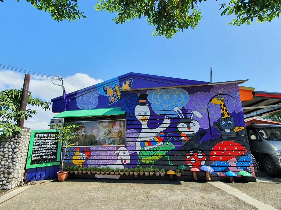A colorful mural of animals and mushrooms decorates the side of a building under a tree, with planters lining the ground.