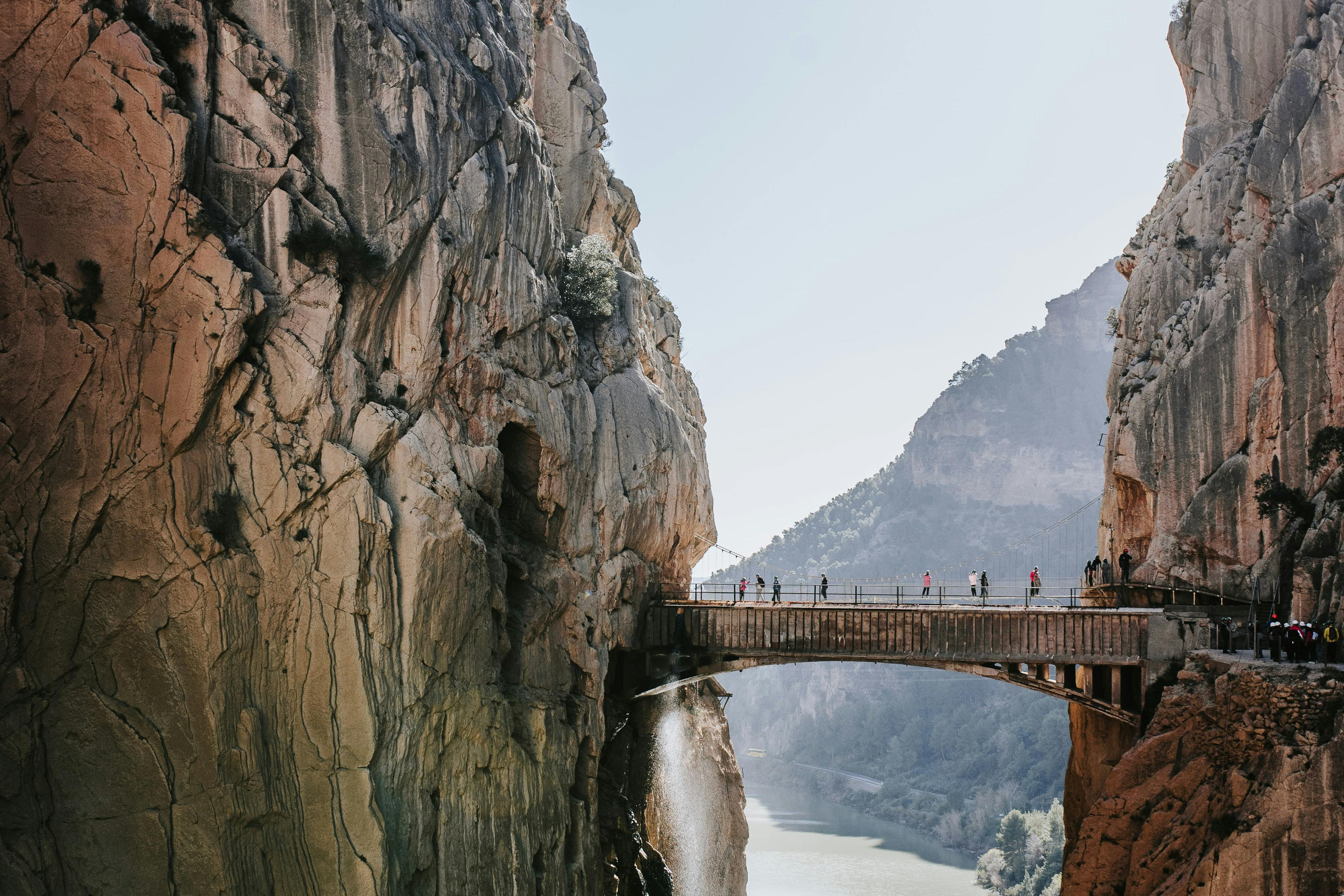 人们走在一座悬在河上陡峭悬崖之间的桥上，背景是山地景观。