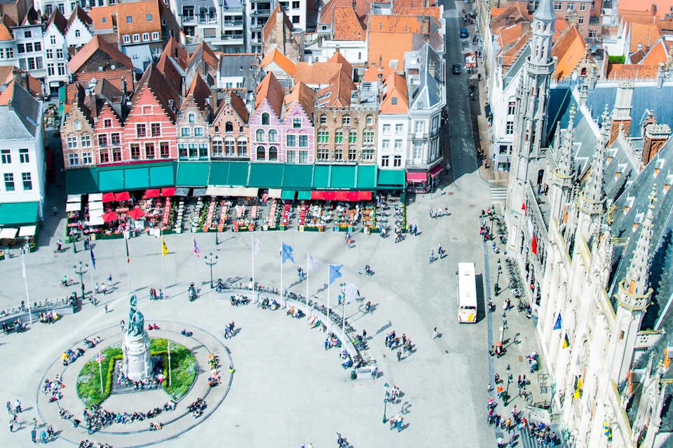 Aerial view of a large, busy square with a statue, flags, and people. Colorful buildings with red roofs and outdoor seating in the background.