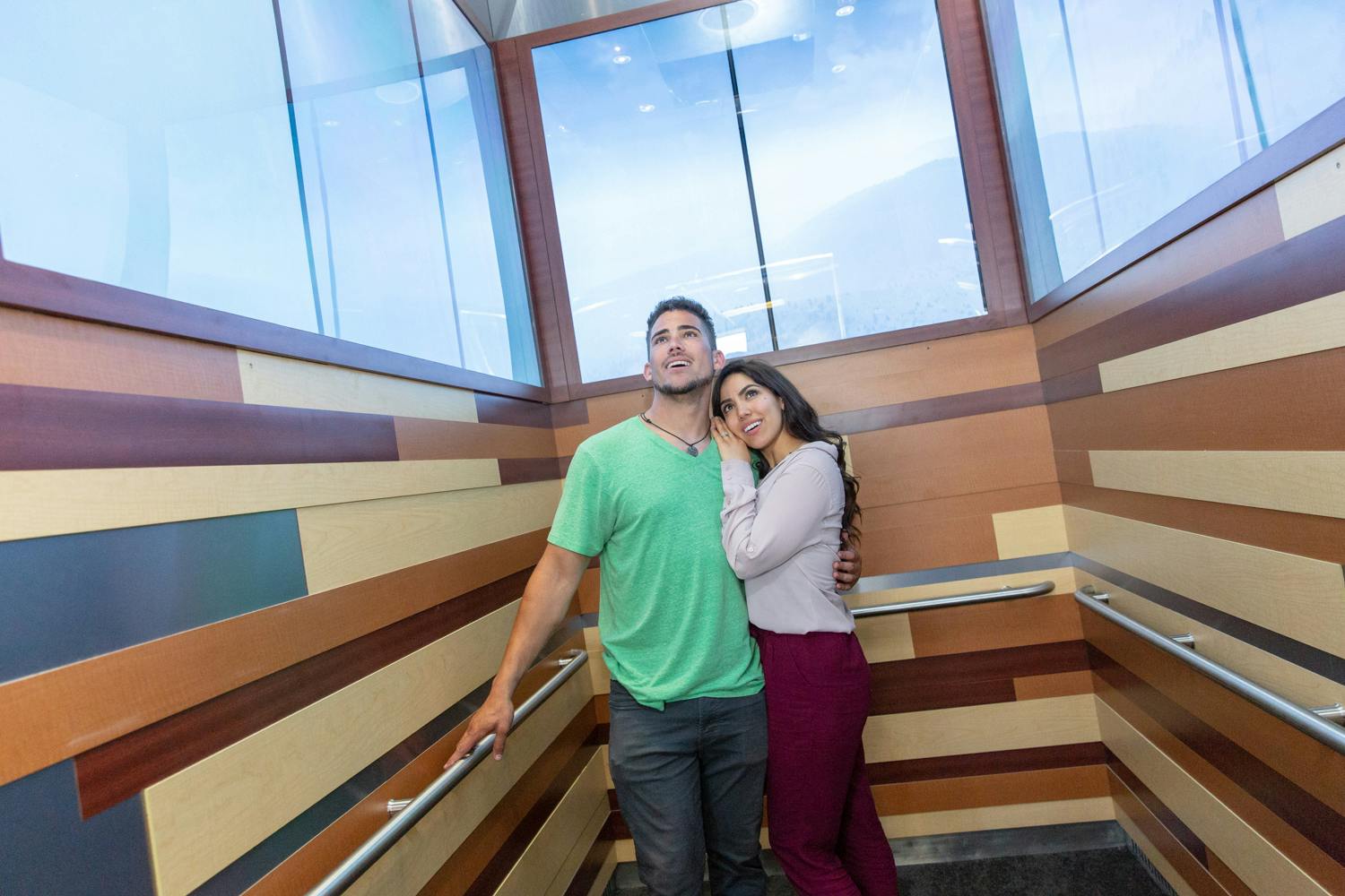 Пара стоит близко друг к другу в лифте с деревянными панелями, обе смотрят вверх с улыбками.