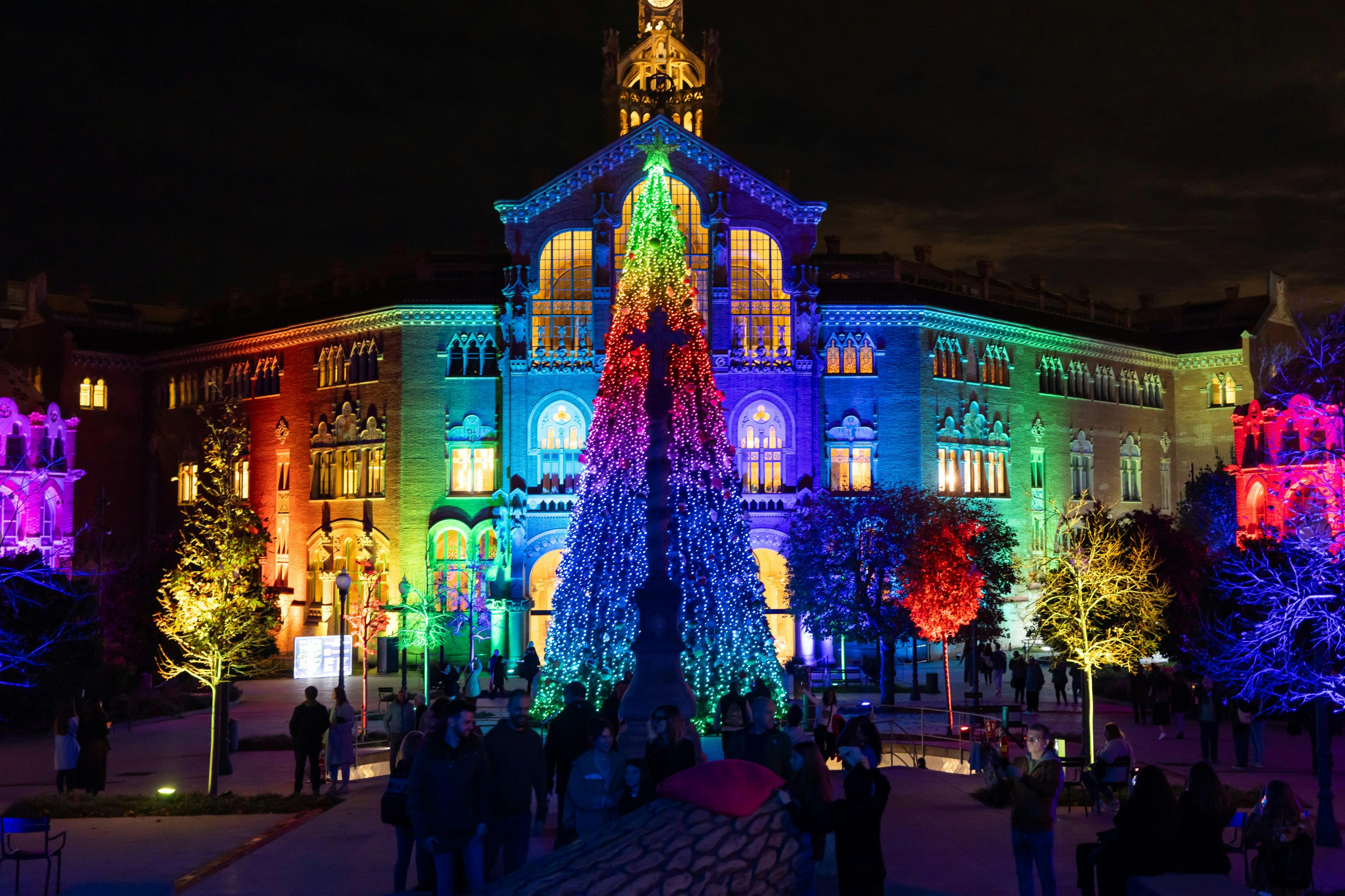 A colorful building illuminated with bright lights and a large Christmas tree surrounded by people at night.