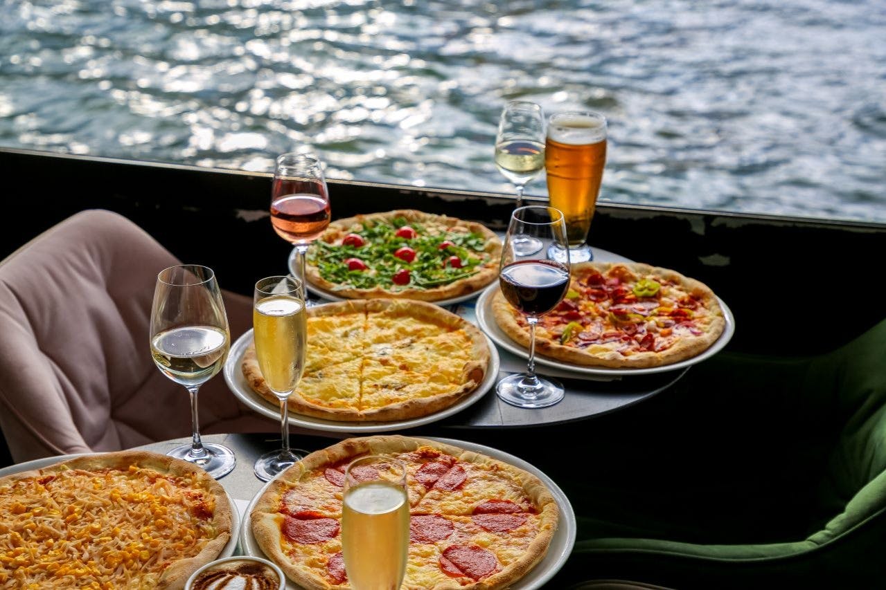 5種類のピザと、ワインやビールなど様々なドリンクが、水をバックにテーブルに並べられています。