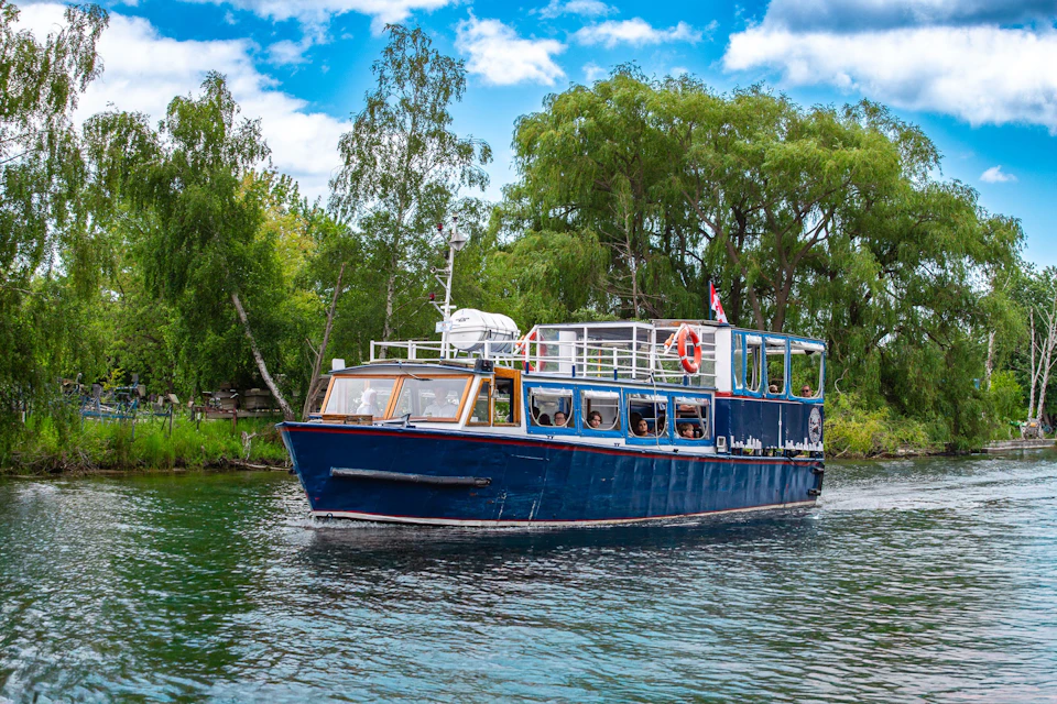 Blue passenger boat cruising on a river, surrounded by lush green trees and a partly cloudy sky overhead.