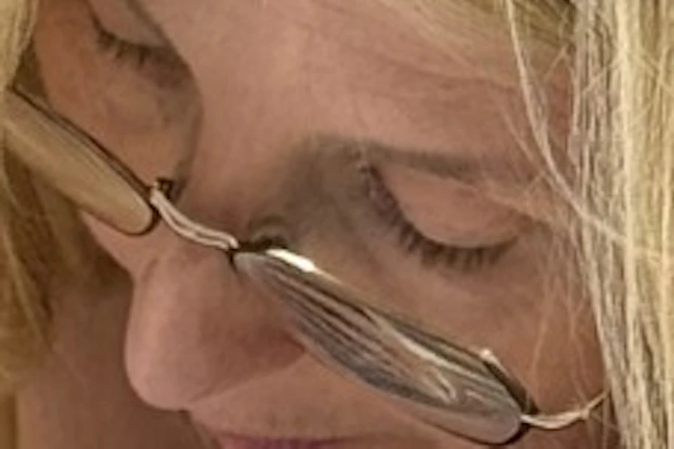 A woman with long blonde hair wearing glasses is painting a decorative pattern on paper at a table with art supplies.
