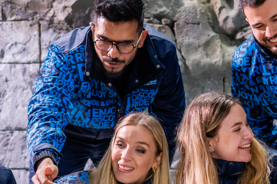 Five people in blue jackets interact with two penguins in a snowy, rocky enclosure.