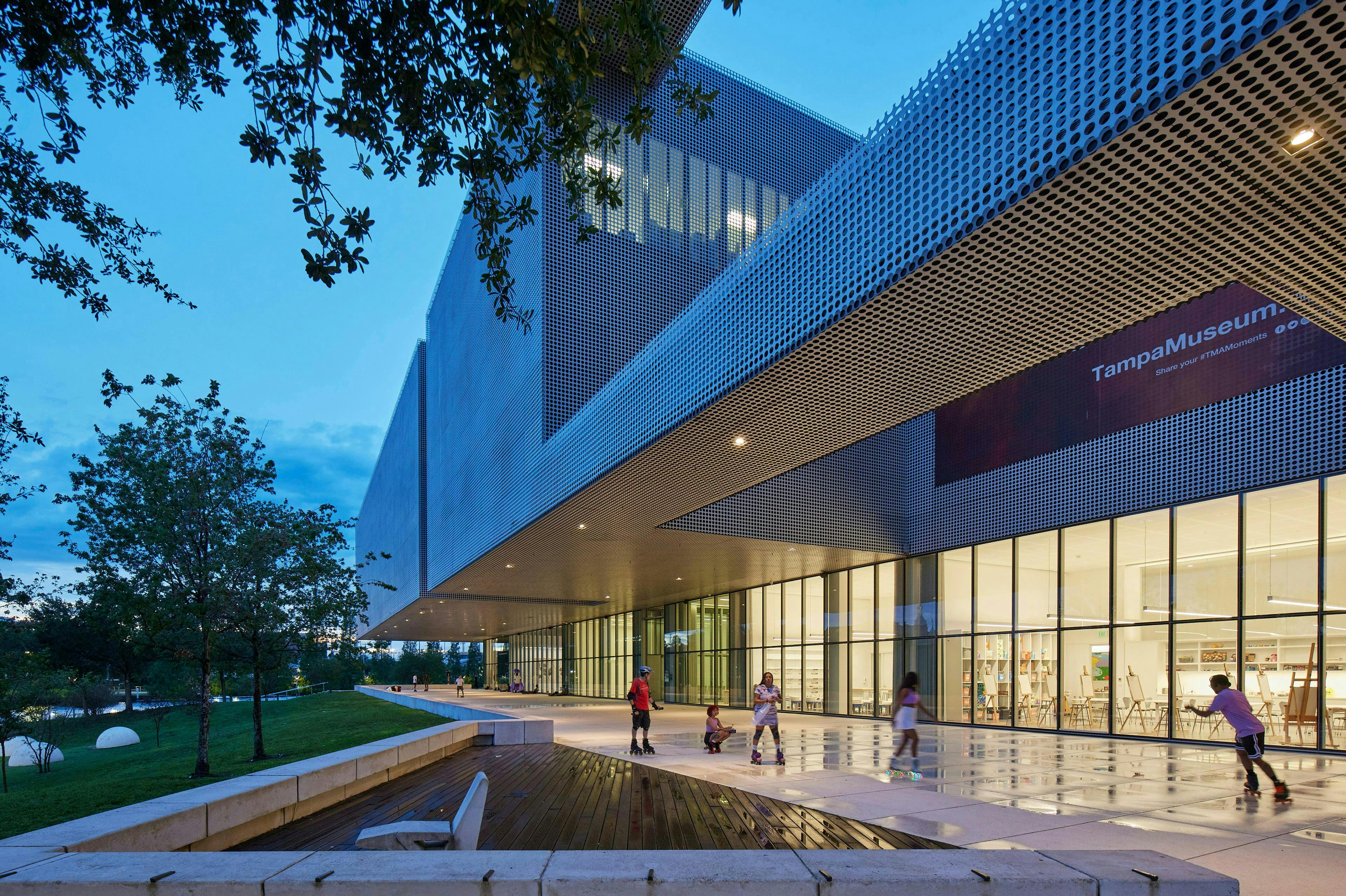 Modernes Gebäude mit perforierter Fassade, großen Fenstern und Rollschuhfahrern in der Nähe in einer abendlichen Umgebung mit Bäumen.