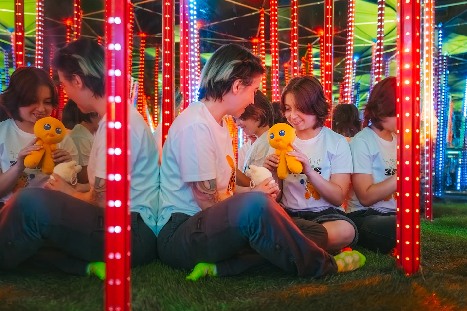 Two people sitting on grass, smiling and holding a yellow plush toy, surrounded by LED-lit pillars and mirrors.