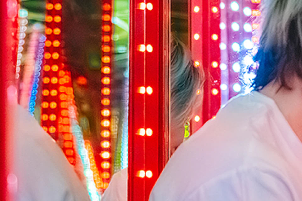 Two people sitting on grass, smiling and holding a yellow plush toy, surrounded by LED-lit pillars and mirrors.