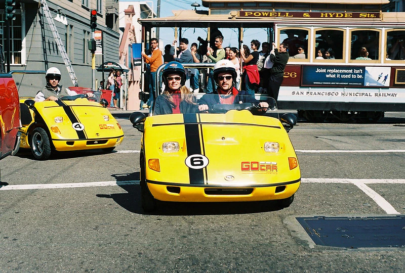Two people in helmets are riding a yellow GoCar on a street. A Powell & Hyde cable car with passengers is in the background.