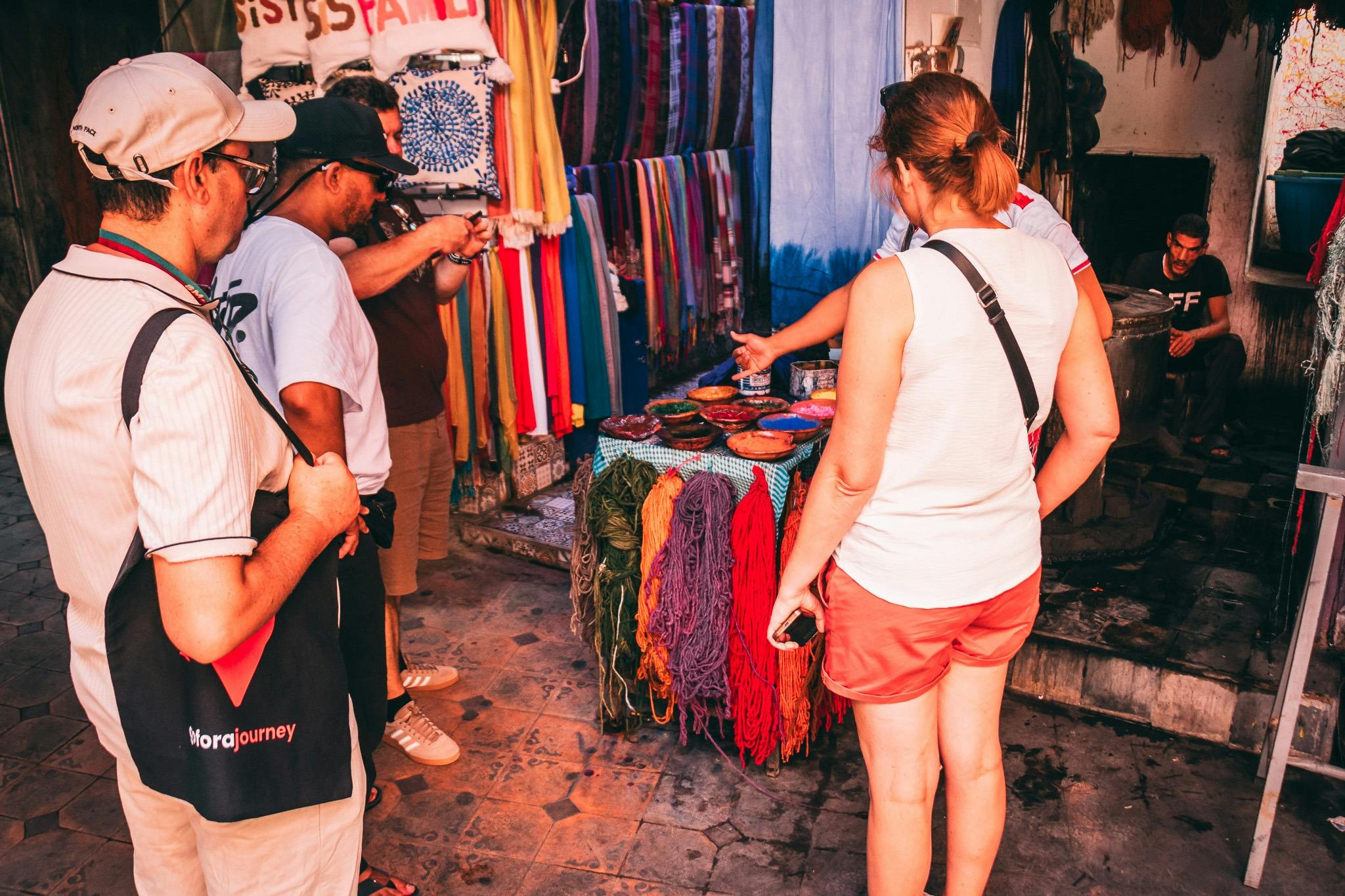 Des gens regardent des tissus colorés et des écheveaux de fil suspendus sur un étal de marché de rue. Une personne prend une photo.