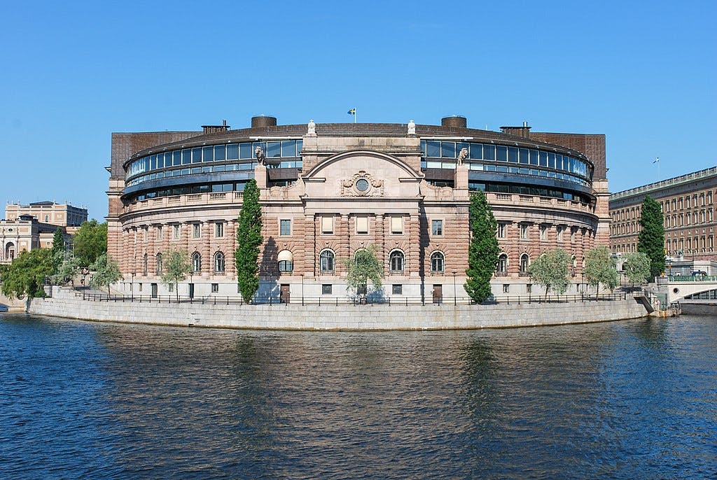 Halvcirkelformad stenbyggnad med välvda fönster och höga träd på flodstranden under en klarblå himmel.