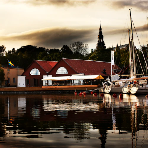 Yachter dockade vid en småbåtshamn med byggnader med röda tak i bakgrunden. Flaggor är synliga och träd omger området.