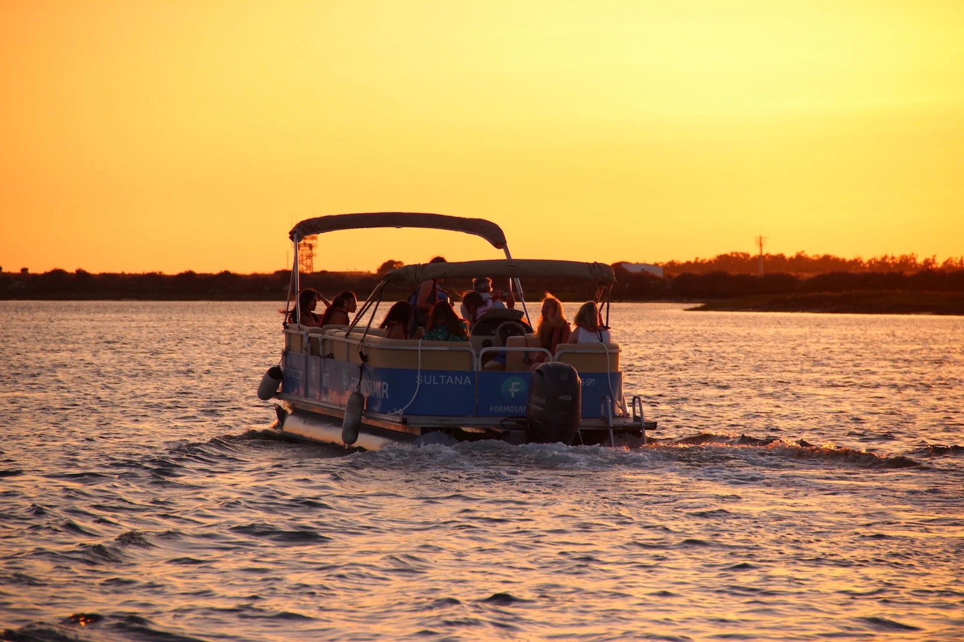 A group of people on a pontoon boat cruising on the water at sunset, with a golden sky and calm water reflecting the light.