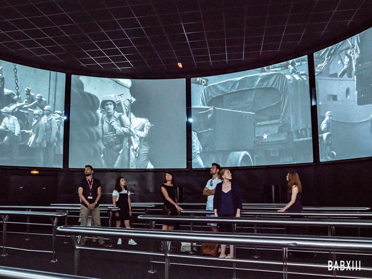 Persone in una stanza buia con grandi schermi a muro che mostrano immagini di guerra in bianco e nero.