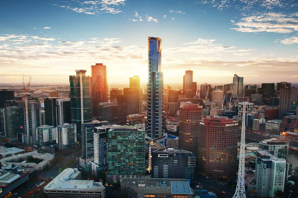 A cityscape at sunset featuring modern high-rise buildings, with a tall blue skyscraper prominently in the center.