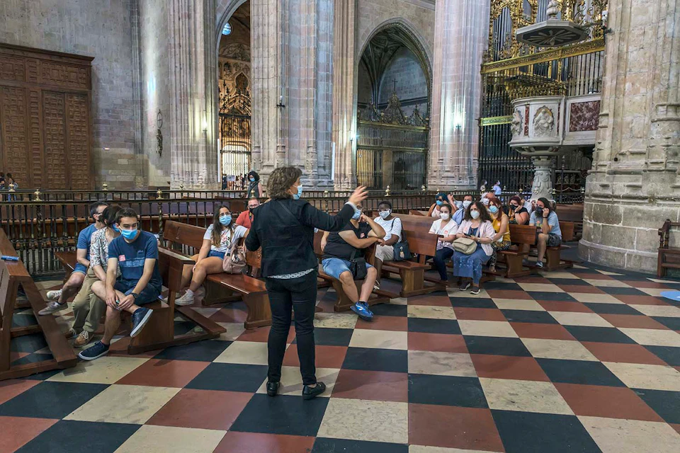 A guide speaks to a group of people seated in a cathedral with tall arches, ornate decor, and checkered flooring.