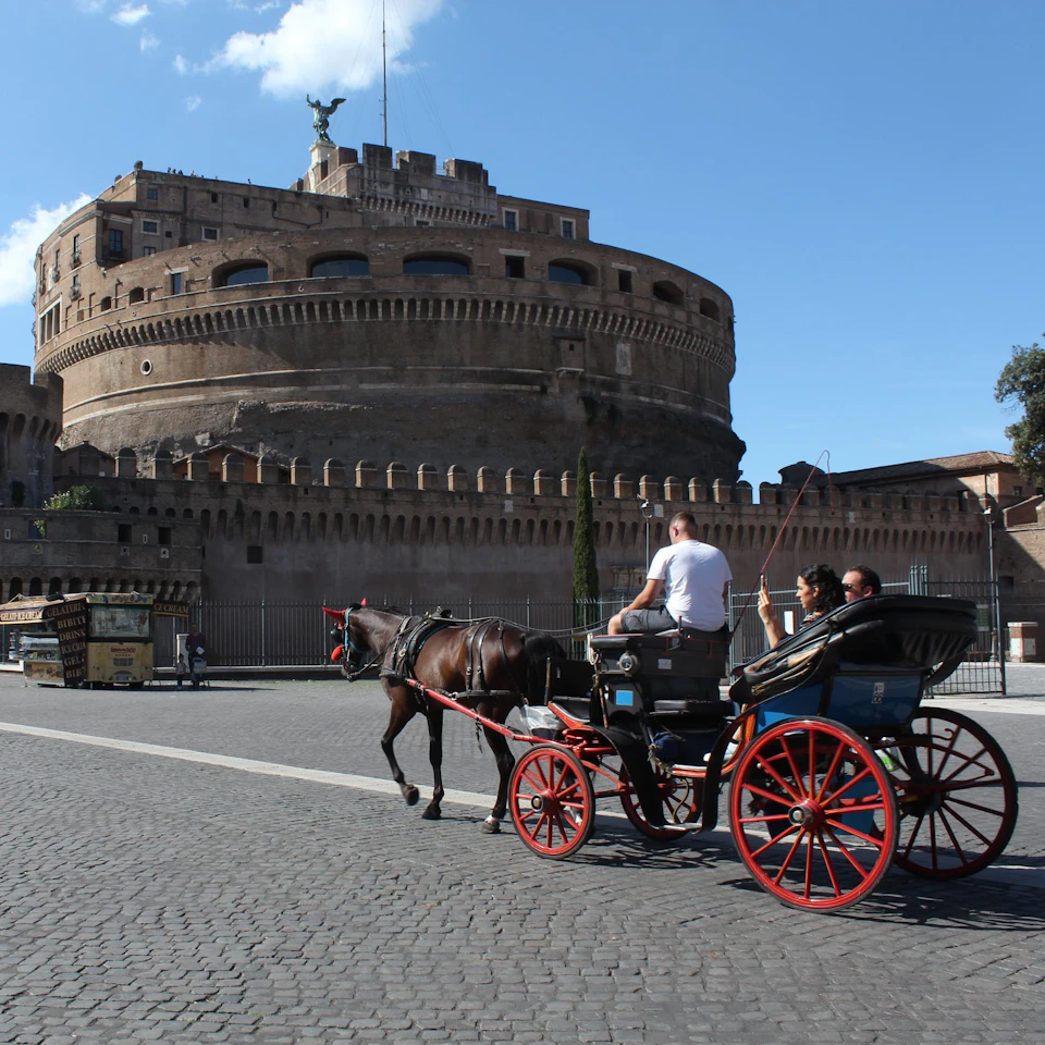 A horse-drawn carriage with three passengers passes by a large, circular stone fortress under a blue sky with few clouds.