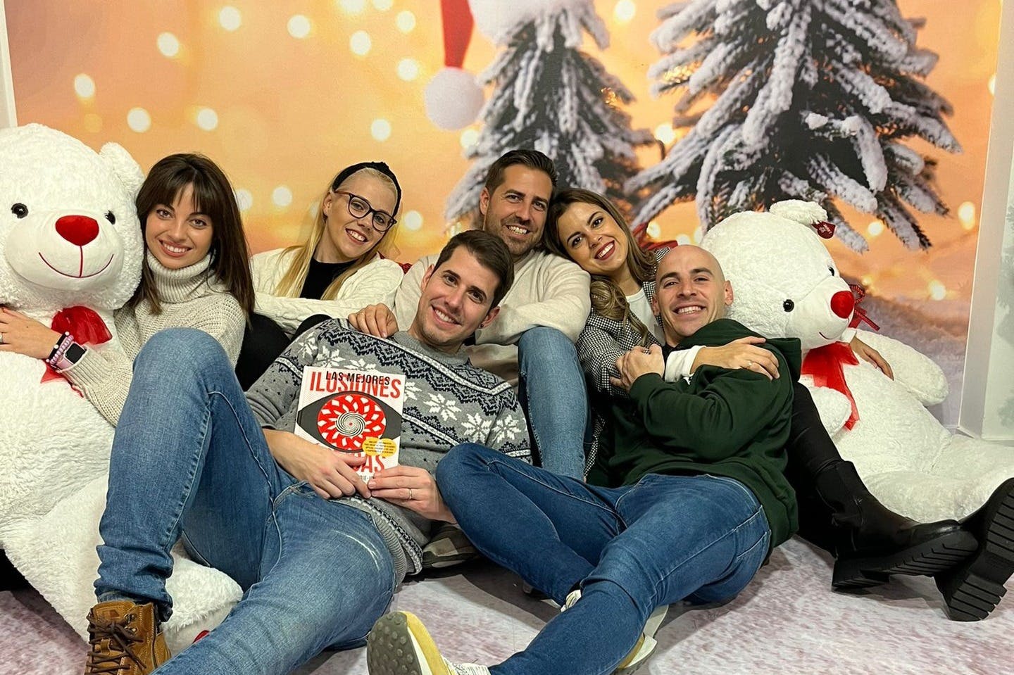 Группа из шести человек, сидящих вместе, улыбаясь, с большим белым плюшевым медведем и праздничными украшениями на заднем плане.