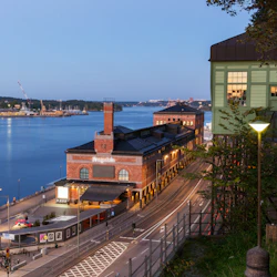 En utsikt över vattnet under skymningen med en historisk tegelbyggnad, en grön byggnad på styltor och en avlägsen stadsbild över vattnet.