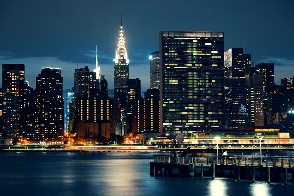 Nighttime cityscape with illuminated skyscrapers reflecting on calm water. A pier and various lit buildings are visible.