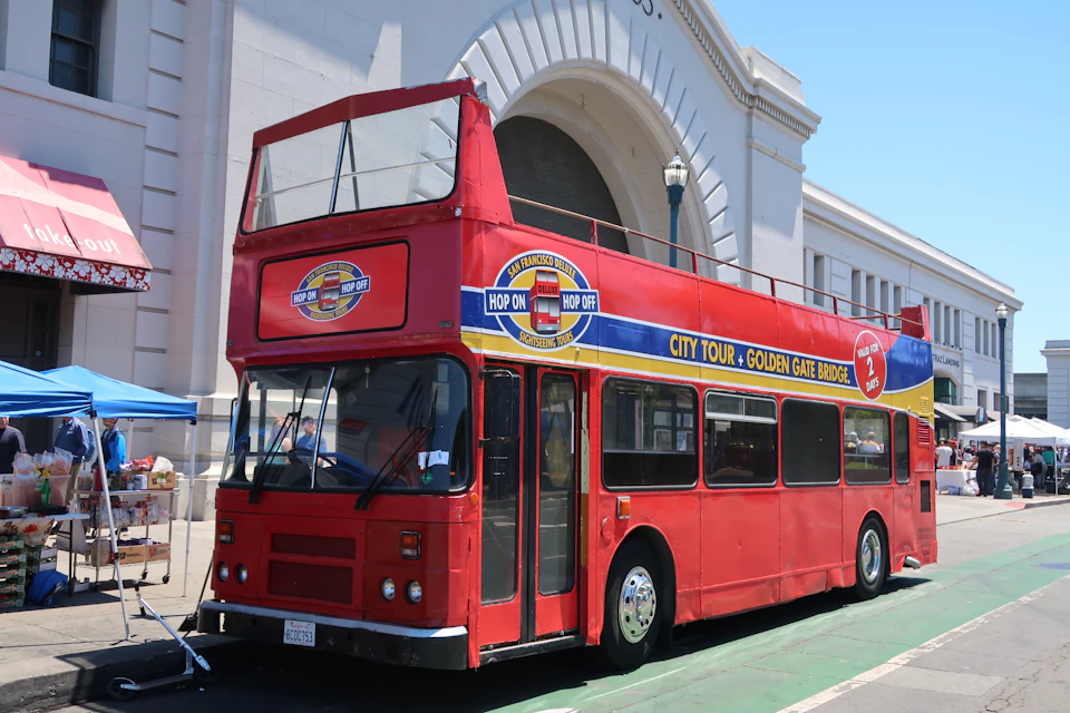 A red double-decker tour bus with a “Hop On Hop Off” sign is parked in front of a large arched building facade.