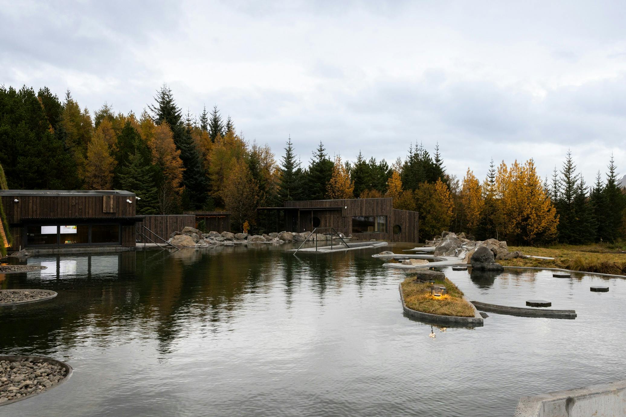 A wooden building by a pond, surrounded by autumn trees and rocks under a cloudy sky.