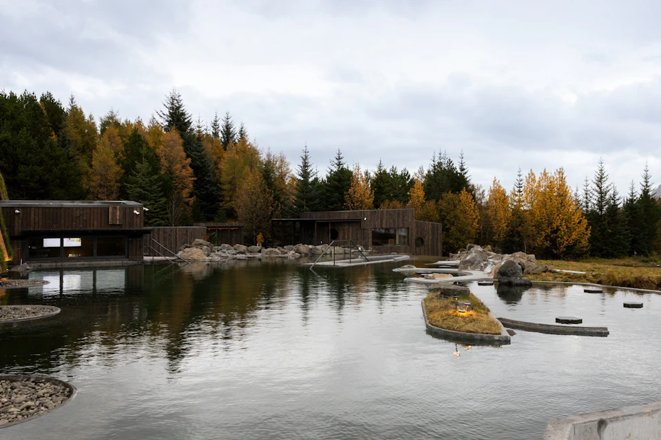 A wooden building by a pond, surrounded by autumn trees and rocks under a cloudy sky.
