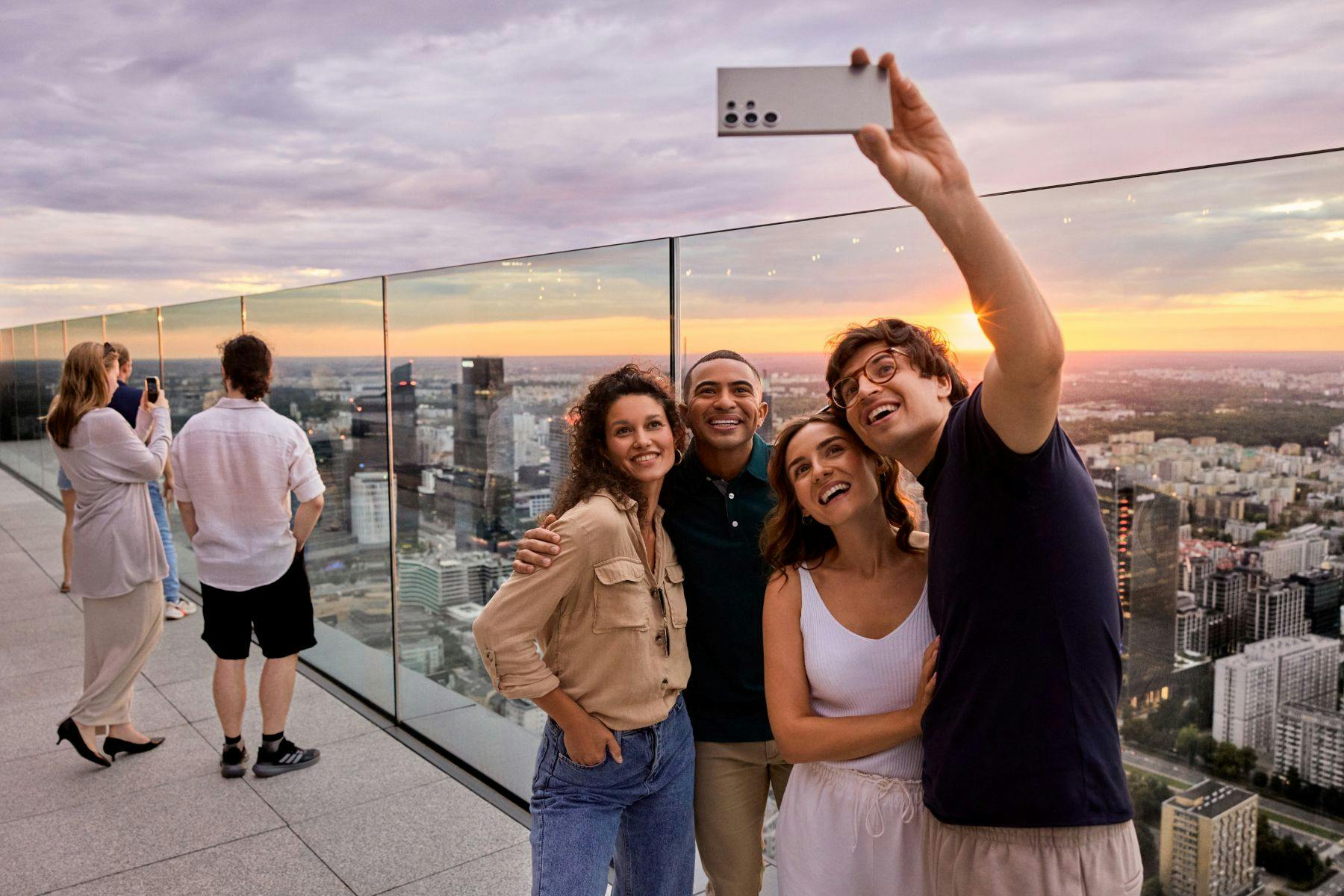 Quatre persones prenent-se una foto en un terrat a la posta de sol, amb un paisatge de la ciutat visible al fons.