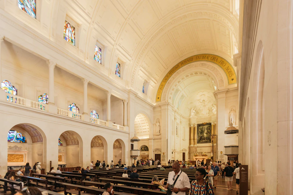 Interior of a large ornate church with people seated and walking. Stained glass windows, high arches, and an altar are visible.