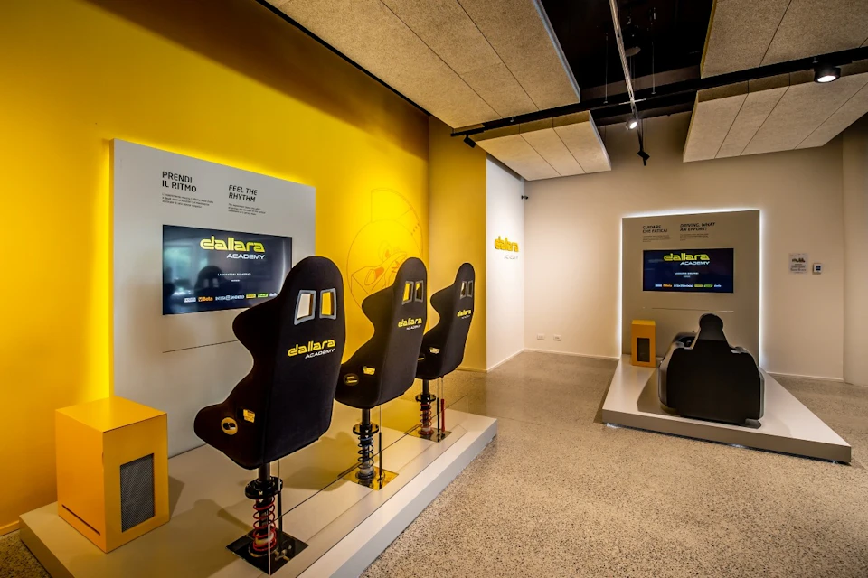 A room with yellow walls featuring three black racing simulator seats, screens displaying "Dallara Academy," and overhead lighting.