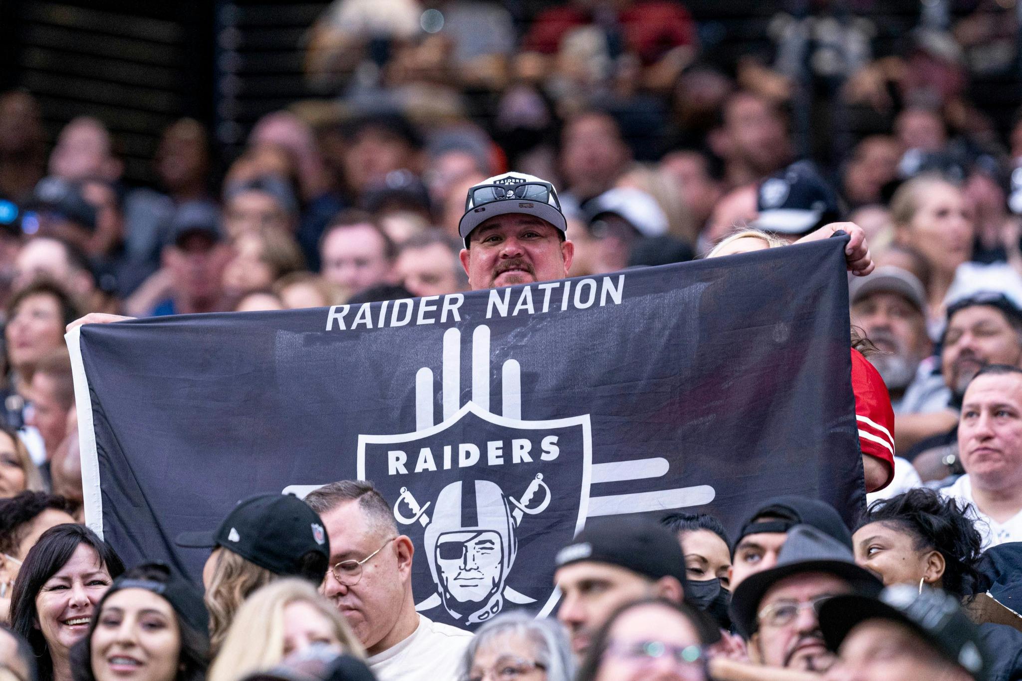 A man in a crowd holds up a Raiders banner with the text 'RAIDER NATION' and the team's logo during an event.
